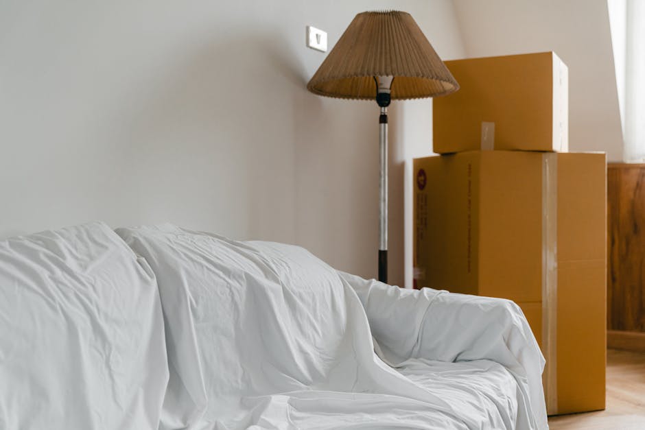 Inside a home used for a house relocation, three stacked cardboard boxes of different sizes sit near a wooden cabinet next to a window, with natural light illuminating the area. A tall, standing lamp with a brown fabric shade is positioned beside the boxes, casting a soft glow. A white fabric-covered sofa is visible in the foreground, with some creases on the upholstery. The scene is part of a typical interior setting where packing and moving preparations are underway, with the boxes prepared for transport. The background features a neutral-colored wall with a small white electrical outlet, emphasizing the indoor environment. This scene relates to furniture transport and packing during the moving process, consistent with the services of Removals Watford, as seen in the context of the costs and logistics involved in home relocation.