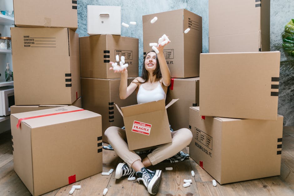 A young woman with long dark hair, sitting cross-legged on a wooden floor inside a room during a home relocation, surrounded by multiple large cardboard moving boxes stacked against the light blue wall. She is wearing a sleeveless white top, beige pants, and striped sneakers, and is smiling while throwing packing foam peanuts into the air. Some boxes are sealed with red tape, and one box exhibits a 'Fragile' label. Behind her, there is a small white shelf with a bottle on it, and to the right, a potted plant is partially visible. The scene captures the packing stage of a furniture transport or moving process, with the woman actively involved in packing or unpacking, and the overall environment suggests an indoor space prepared for a house move in Watford, with natural lighting illuminating the area. Removals Watford offer professional services related to packing and moving, as illustrated by the scene of packing and handling boxes in the home.